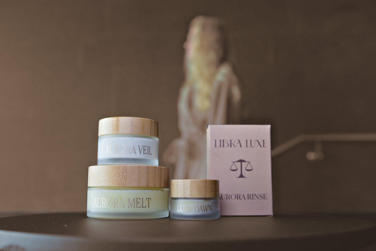 Aurora Veil face cream, Aurora Melt cleansing balm and Luna Dawn lip mask beside the Aurora Rinse solid cleanser box on a dark table, a woman out of focus behind them