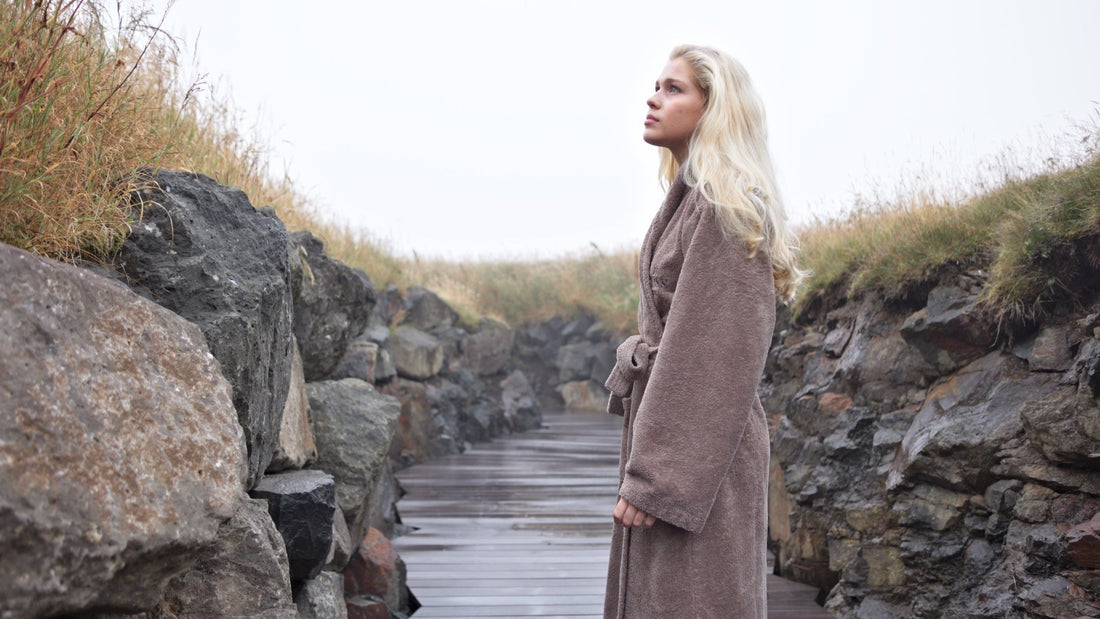 Woman in a taupe bathrobe on a wet boardwalk between stone walls and tall grass, looking up into the fog, Faroe Islands